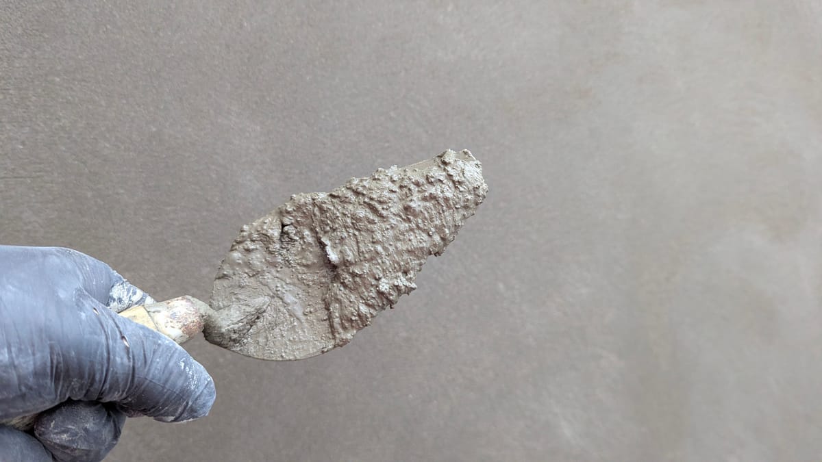 Hand im Arbeitshandschuh mit einer Kelle voller Putz vor einer glatten grauen Wand
